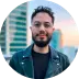 Portrait of a man with glasses and a beard, wearing a dark leather jacket and dark t-shirt. He is in front of a city background, looking at the camera with a small smile. He has dark, curly hair