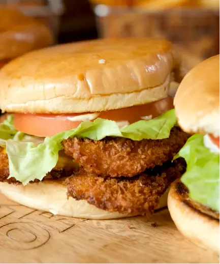 A close-up photo of a crispy chicken sandwich. The sandwich has two breaded and fried chicken patties, lettuce, tomato, and mayonnaise on a toasted bun. There is another burger partially visible in the background