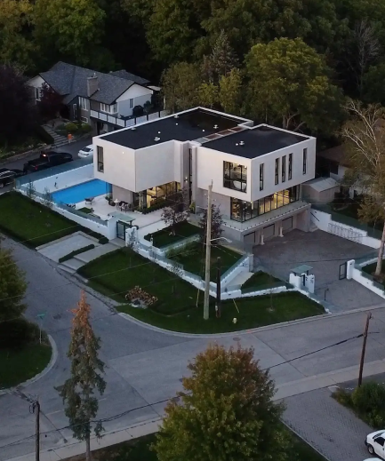 Aerial view of a modern, two-story house with a flat roof. The house is surrounded by trees and has a swimming pool in the backyard. There is a driveway leading up to the house and several cars parked in the driveway