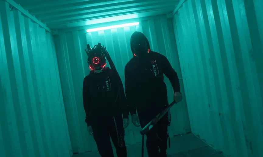 Two hooded figures in a dark, neon-lit hallway. One figure is wearing a mask with glowing red eyes and the other is carrying a baseball bat