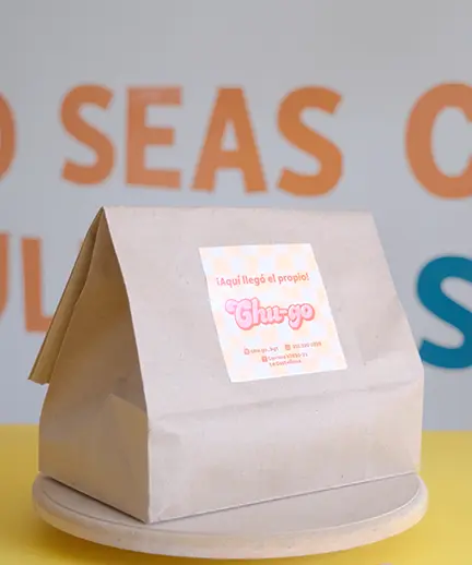 A brown paper bag with a label featuring Spanish text. The background displays a sign with orange and blue lettering