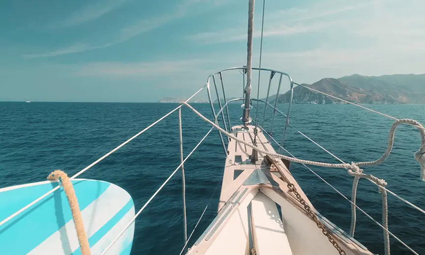 A sailboat sailing on a calm blue ocean. The bow of the boat is visible, with ropes and a metal railing. A surfboard is tied to the side. In the distance, there are mountains on the horizon