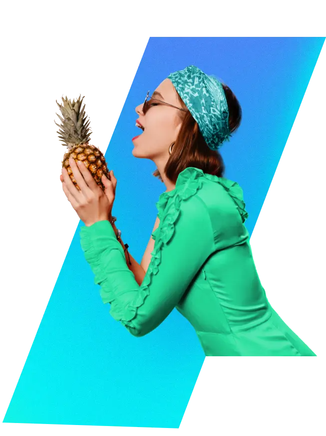 Woman with light brown hair, wearing a green dress and headband, holding a pineapple close to her open mouth in an expression of excitement. She is seen from the side, wearing glasses with her eyes closed